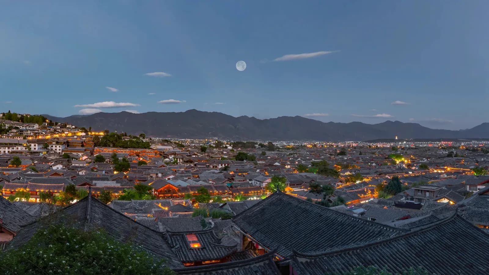 Night rooftops of Lijiang Old Town under a full moon, lanterns glowing toward the mountains