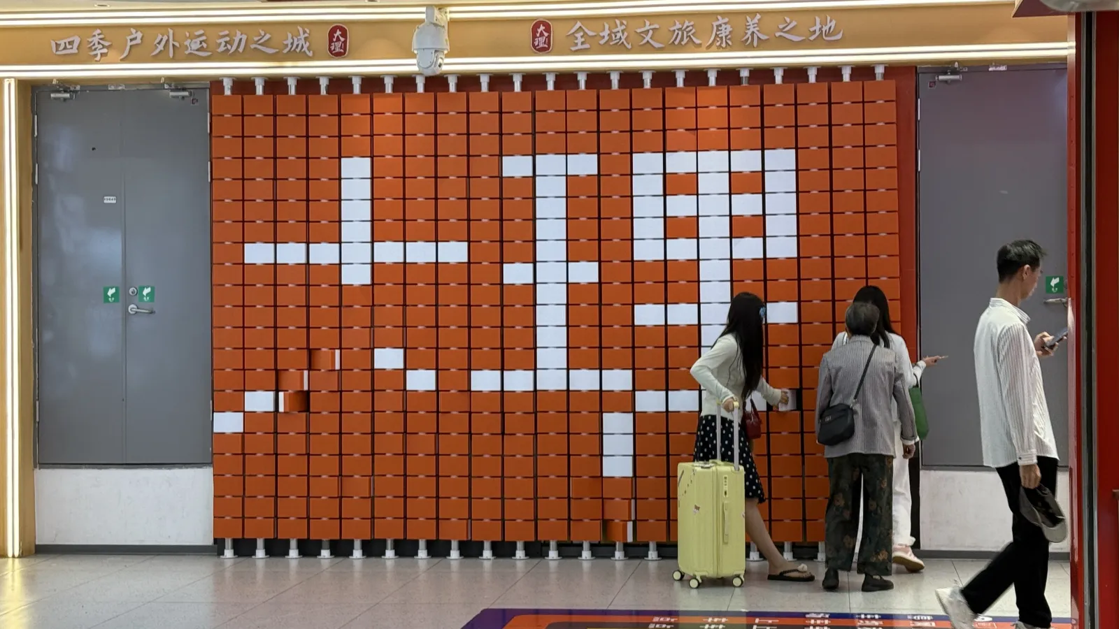 Arrival at Dali railway station Orange block wall spelling Dali in Chinese characters inside the railway station, with travelers and luggage in the foreground