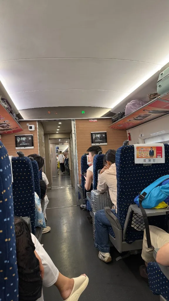 Inside a China high-speed rail carriage Second-class carriage interior on a China high-speed train, with blue seats, overhead luggage racks, and the aisle toward the next car